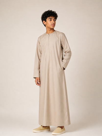 Boy wearing a premium Omani thobe in sand beige with traditional neckline and front zip detail, front view on a neutral studio background.